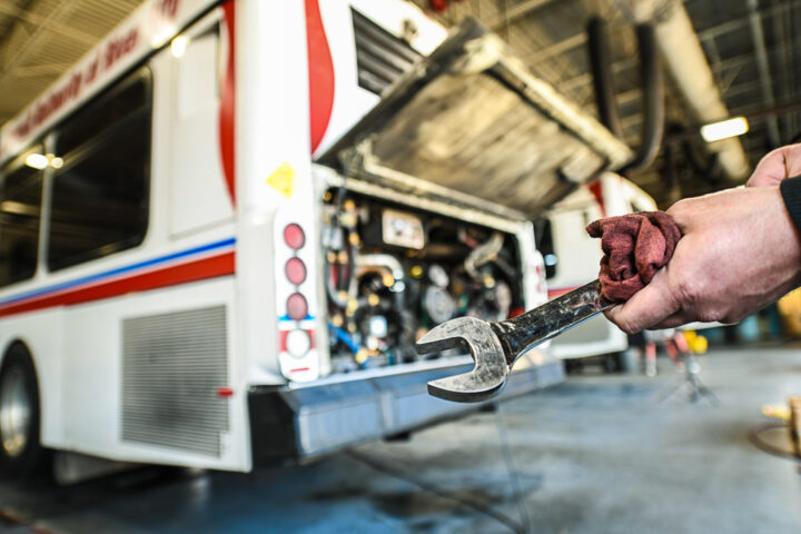 A mechanic holds a wrench and rag in front of a city bus with its engine compartment open inside a maintenance garage.