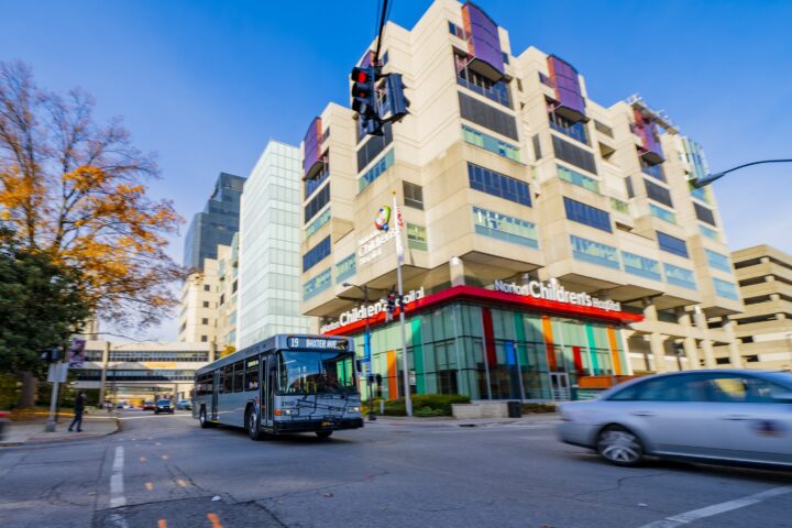 A city bus drives past Norton Children's Hospital on a clear day, with cars and a modern hospital building in the background.