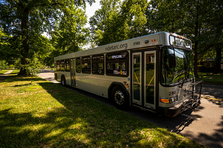 A city bus parked on a shaded residential street with green trees and grass on a sunny day.