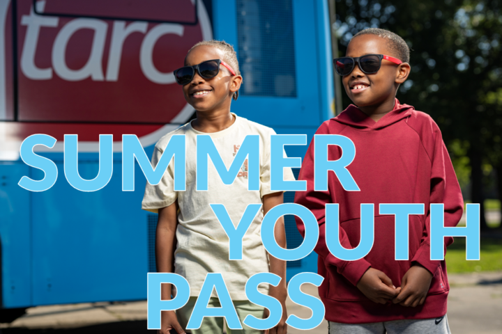 Two children wearing sunglasses stand in front of a blue TARC bus with the words 