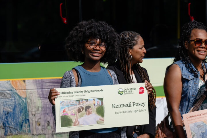 A smiling student holds a poster with her name, Kennedi Posey, and artwork for the Olmsted Parks and TARC's 
