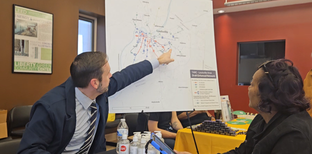 A man points to a location on a large map while speaking to a woman seated beside him at a table in a meeting room.