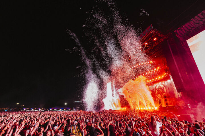 A large crowd at an outdoor concert at night with bright red stage lights, smoke, and confetti filling the air.