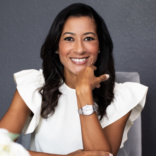 Woman with long dark hair sits at a table, smiling, wearing a white ruffled sleeve top and a silver watch, against a gray background.