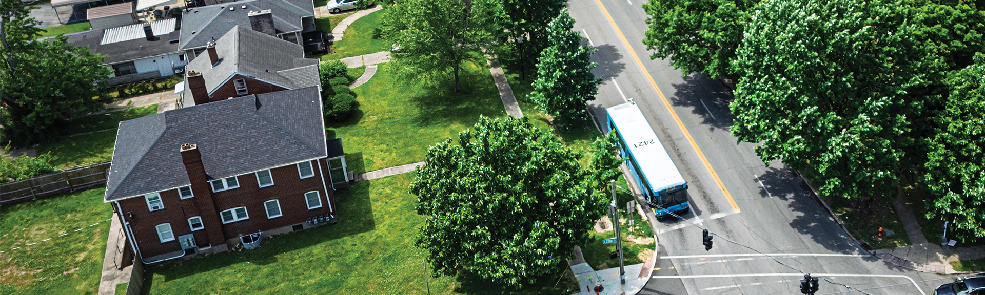 Aerial view of a residential street with a brick house, green lawns, trees, and a city bus stopped near an intersection.